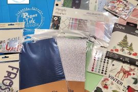 Flat lay photo of The Paper & Ink Boutique Winter Crafting Giveaway prize package featuring Simple Stories Flannel Season collection, Prism Studio cardstock, Sizzix and 49 & Market stamp sets, embellishments, and a blue Paper & Ink Boutique tote bag.