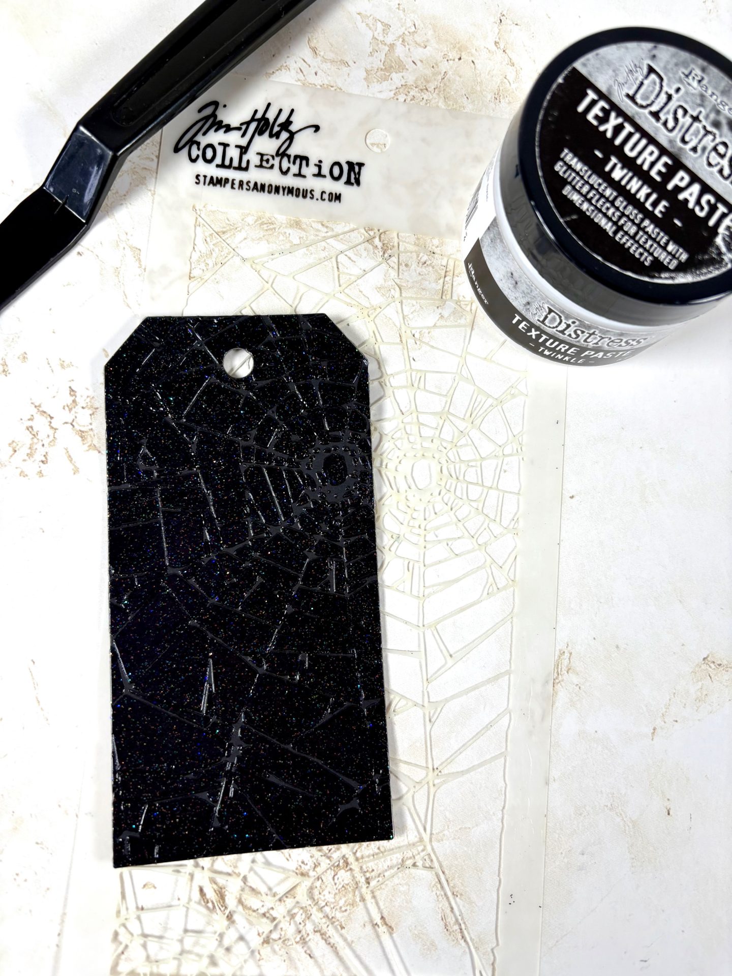 Step 3 of Spooky Treat Tags project by Heather Fischer showing a black die-cut tag made with Ranger Black Mixed Media cardstock, decorated using Stampers Anonymous THS014 Shatter stencil and Ranger Distress Texture Paste Twinkle for a glittery cobweb effect.
